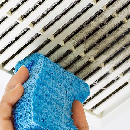 Hand cleaning an air vent grille using a blue sponge as part of air duct maintenance.