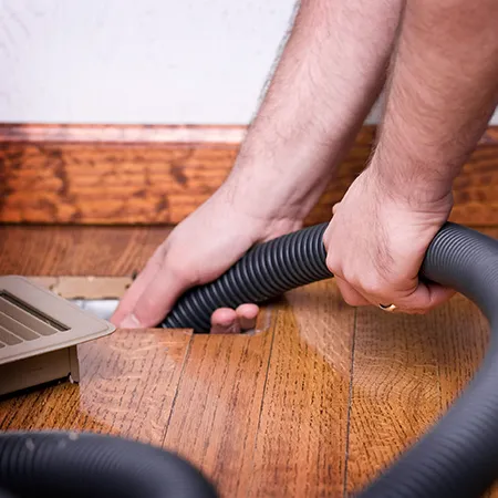 Hands using vacuum hose to clean air duct floor vent during professional air duct sanitization in Brookfield WI.