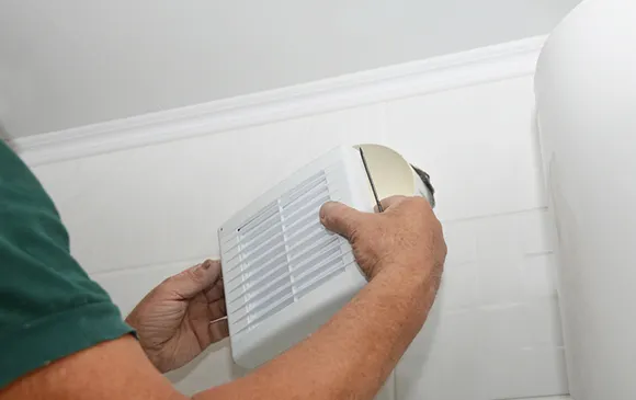 Person cleaning a white bathroom exhaust fan grille mounted on ceiling for better ventilation.