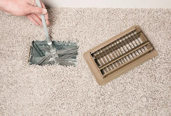 Person cleaning floor vent with vacuum brush attachment to remove dust and debris.