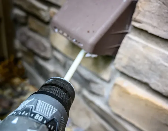 Power drill fixing a metal dryer vent cap attached to a stone wall in outdoor dryer vent repair.