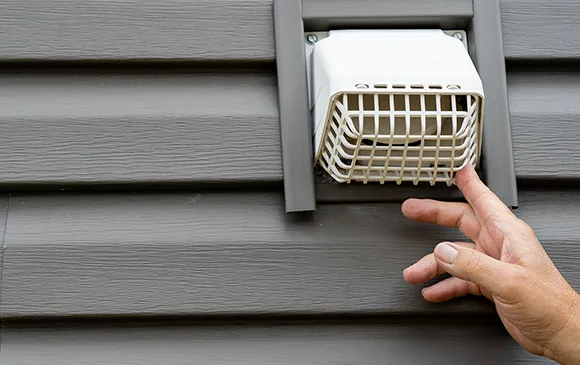 Person removing or inspecting exterior dryer vent cover on house siding.