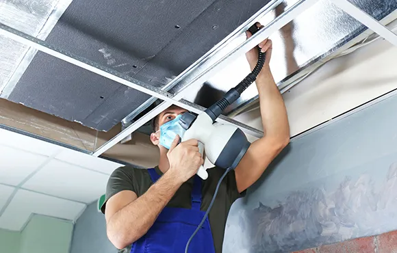 HVAC technician in protective gear cleaning and sanitizing overhead air ducts for thorough air duct sanitization service.