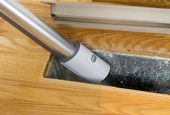 Vacuum hose cleaning dust and debris from hardwood floor air duct vent.